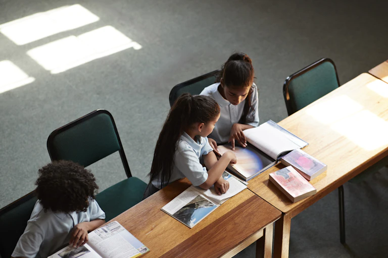 Three students seated at a long desk read books.