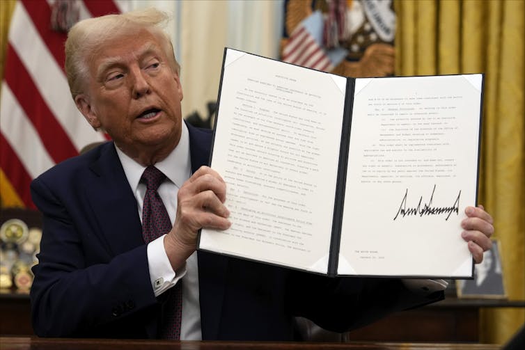 A white man with orange-tinged skin and white hair holds up a signed document in a portfolio
