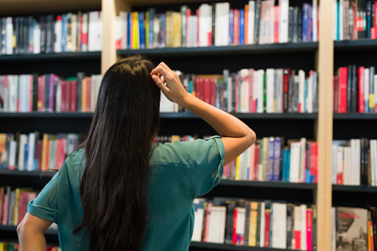 A young woman, seen from behind, scratches her head while looking at shelves of books
