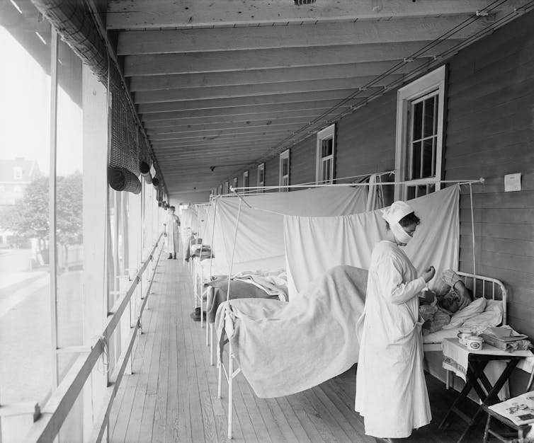 Black and white photo of a hospital ward with beds separated by whtie sheets and a nurse in the foreground