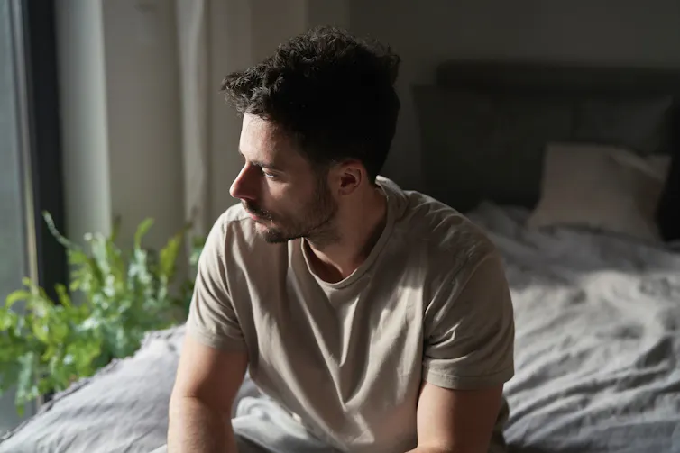 A young man sitting on a bed, appears pensive.