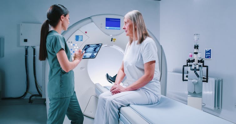 Health worker talking to older woman sitting on bed of MRI scanner.