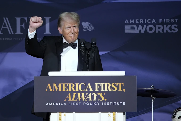 US President Donald Trump standing with raised fist behind a podium reading "America First, Always".