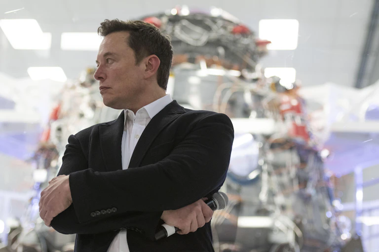 Elon Musk at SpaceX headquarters, with his arms crossed.