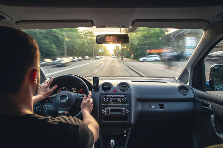 A man sits in the driver's seat of a car. His hands are both on the wheel, and part of his face is visible in the rearview mirror.