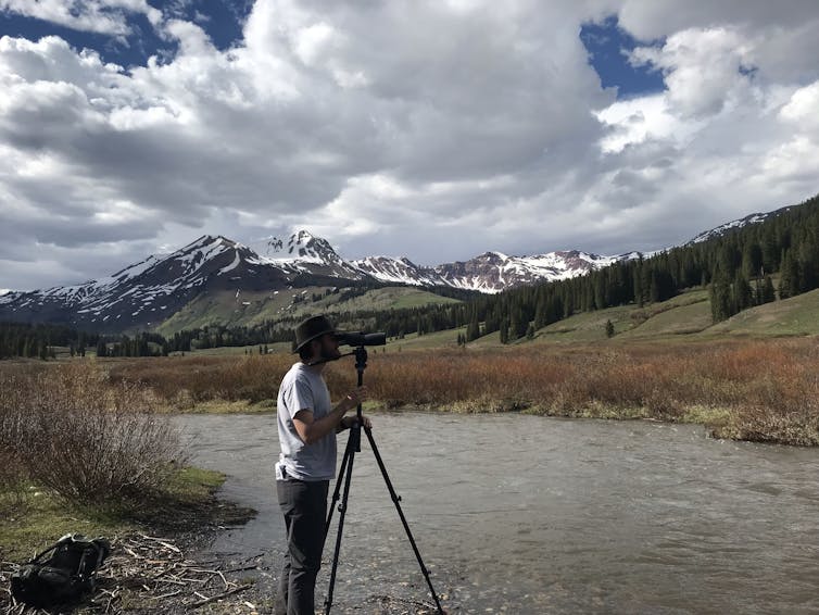 man at tripod pointed across river with mountains in background