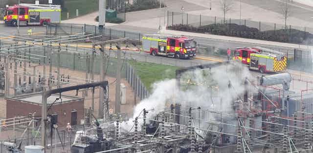 Smoke rising from large installation of electrical equipment with fire engines nearby