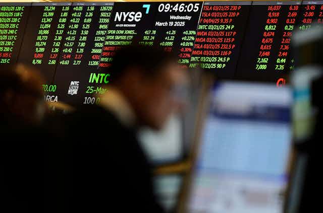 Stock market numbers displayed on a screen behind the blurred image of a person working on the options floor