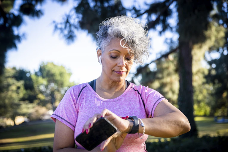 A woman with short white hair standing outside looks at her watch while holding a phone