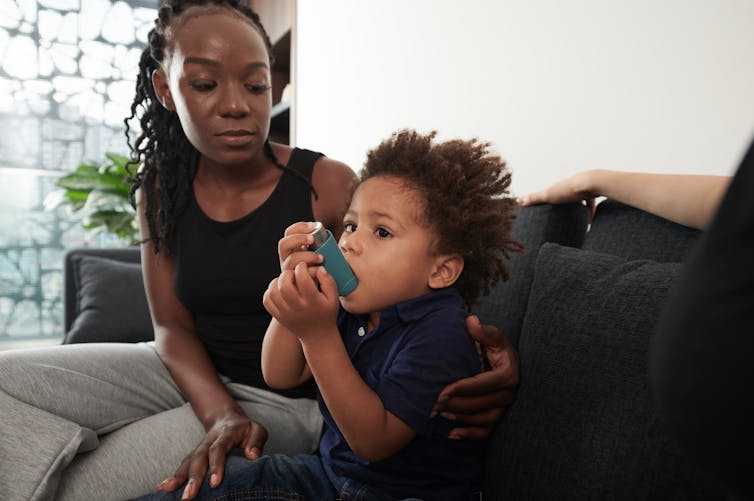 A young toddler uses an asthma inhaler. His mother has an arm wrapped around him while she watches.