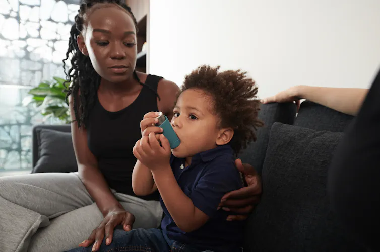 A young toddler uses an asthma inhaler. His mother has an arm wrapped around him while she watches.