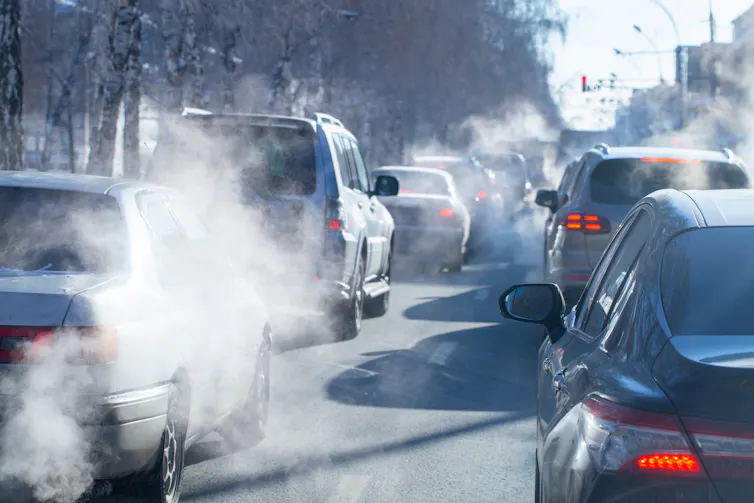 Two rows of cars idle on the road, with pollution coming out of their exhaust pipes.
