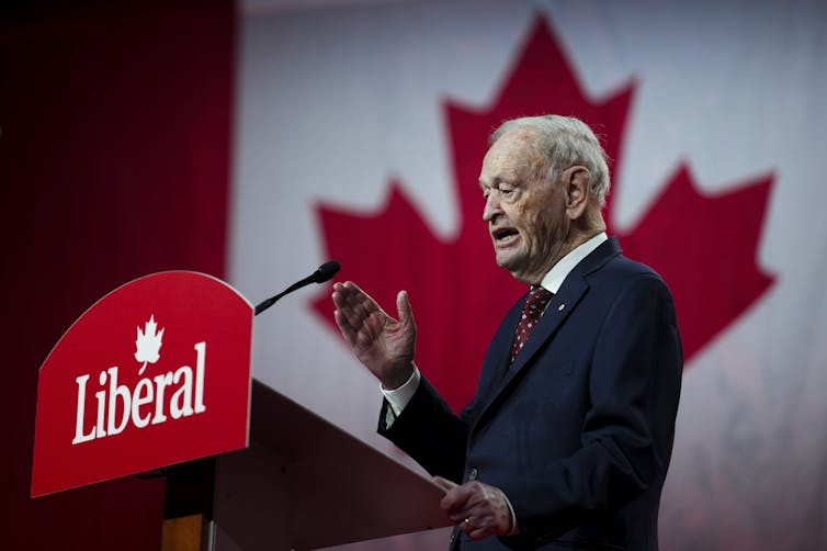 El hombre anciano habla en un micrófono con una bandera canadiense gigante detrás de él.