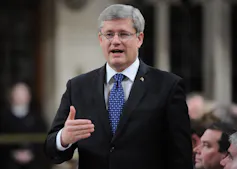 A grey-haired man in a dark suit and blue tie and glasses speaks in the House of Commons.