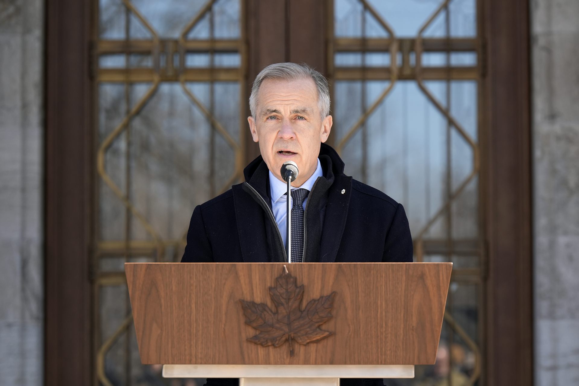 A man with short grey hair speaks into a microphone behind a wooden lectern with a carved maple leaf.