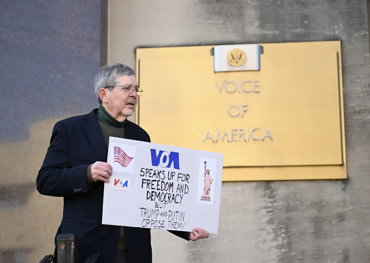 Staff member with sign protesting in front of Voice of America sign.