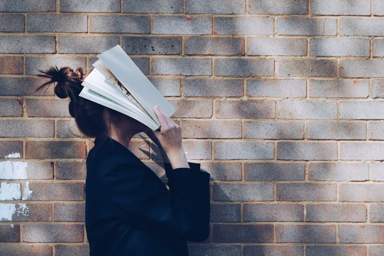 A woman in front of a brick wall slamming a book against her face in apparent frustration.