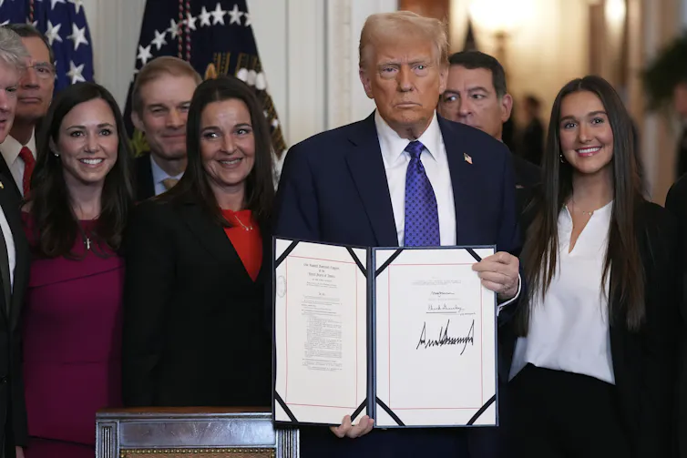 An older man with fluffy white-blond hair holds up a document he's signed.