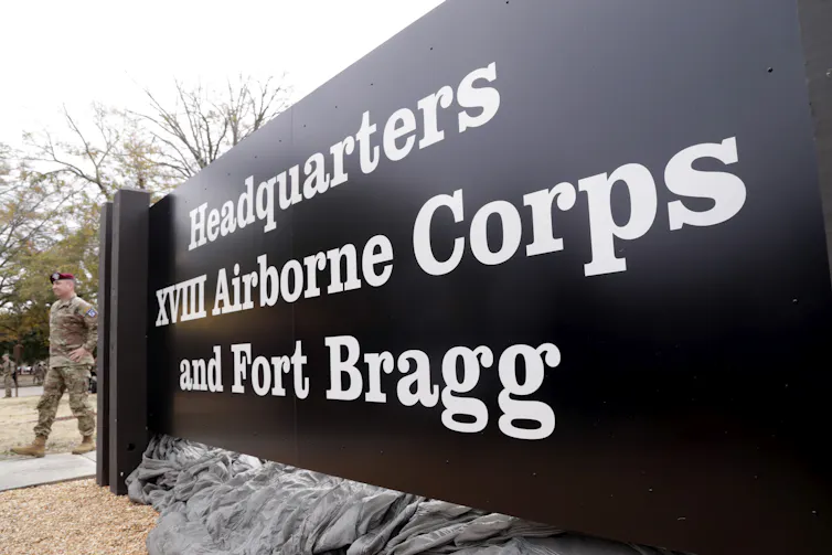 A soldier walks behind a sign reading 'Headquarters XVIII Airborne Corps and Fort Bragg'