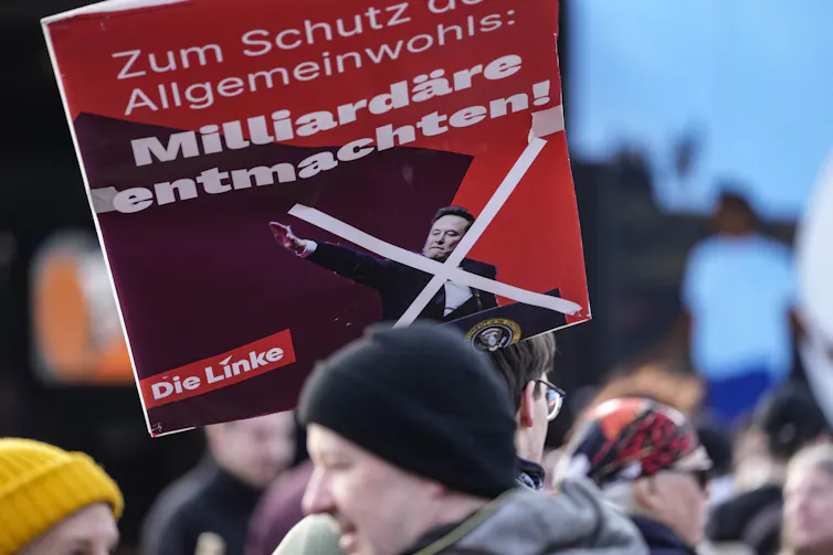 a man wearing a black beanie holds a red sign with a photo of Elon Musk giving a Nazi salute crossed out