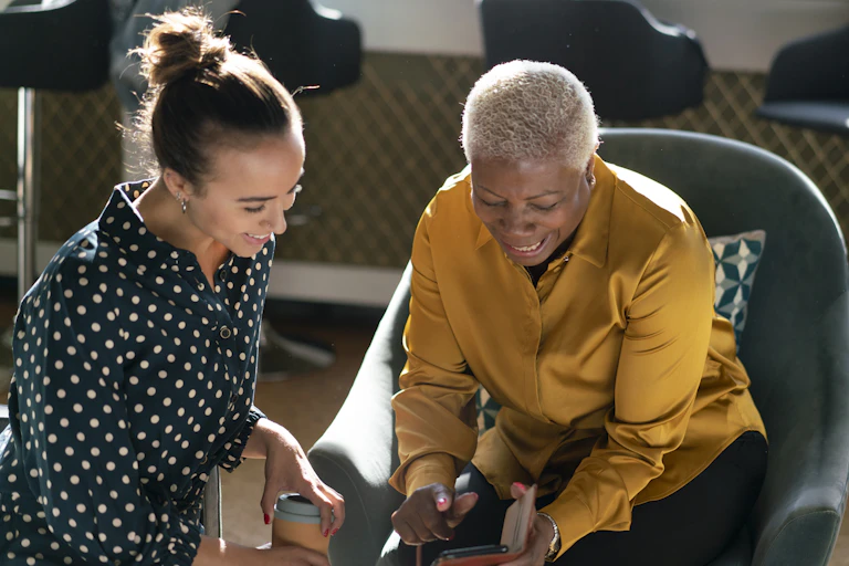 A young woman with brunette hair and an older woman with short gray hair sit as they chat and smile, looking at a phone.