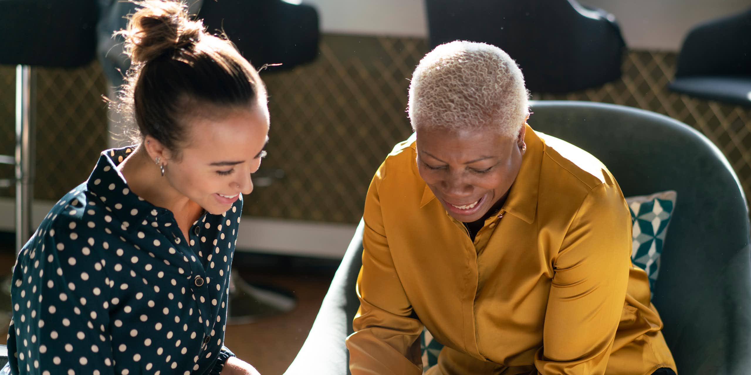 A young woman with brunette hair and an older woman with short gray hair sit as they chat and smile, looking at a phone.