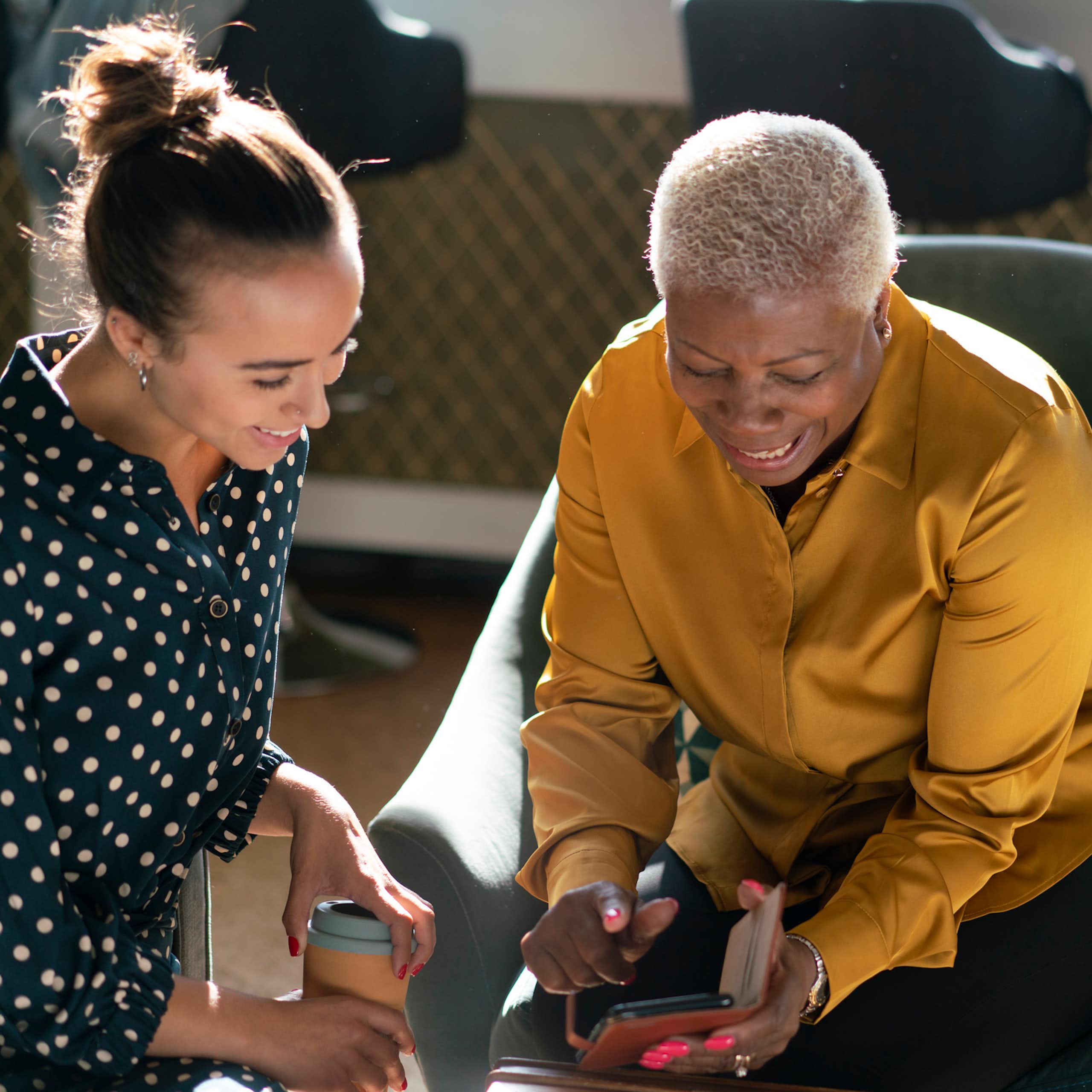 A young woman with brunette hair and an older woman with short gray hair sit as they chat and smile, looking at a phone.