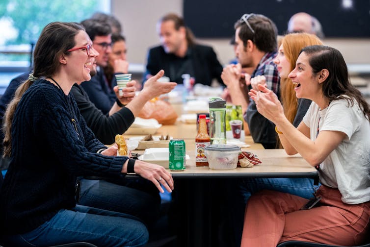 Two young women sit at the end of a long table of young people with food containers and cups in front of them.