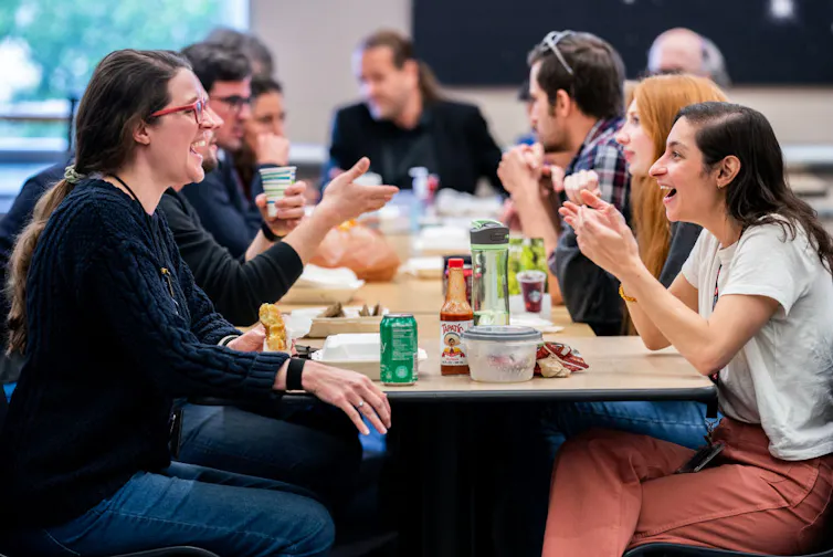 Two young women sit at the end of a long table of young people with food containers and cups in front of them.