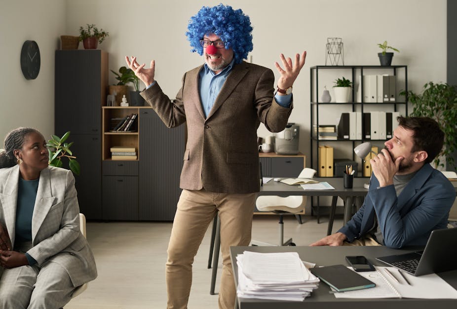 un homme avec une perruque et un nez rouge dans un bureau. les personnes assises semblent consternées.