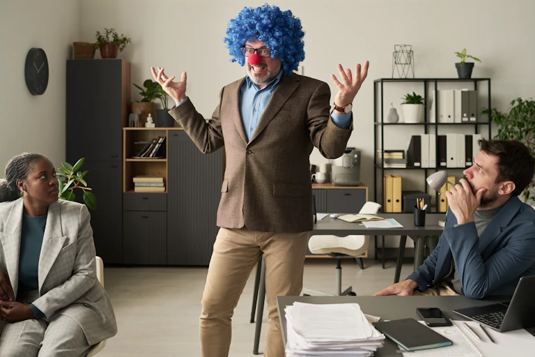 un homme avec une perruque et un nez rouge dans un bureau. les personnes assises semblent consternées.