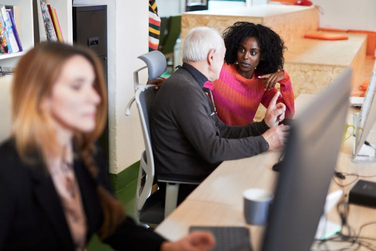 A young Black woman in a pink sweater turns to talk to a white man with white hair in a black turtleneck as they sit in front of laptops.