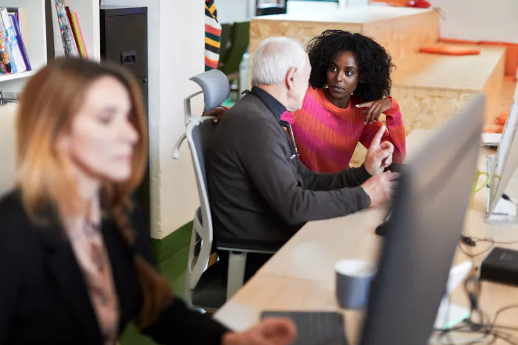 A young Black woman in a pink sweater turns to talk to a white man with white hair in a black turtleneck as they sit in front of laptops.