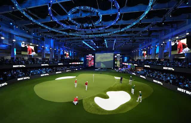 People on an indoor golf green surrounded by stands with an audience