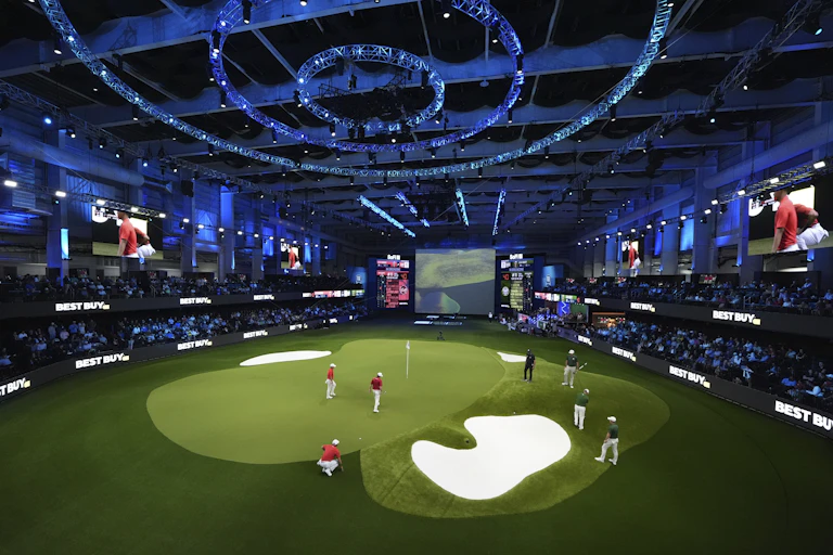 People on an indoor golf green surrounded by stands with an audience