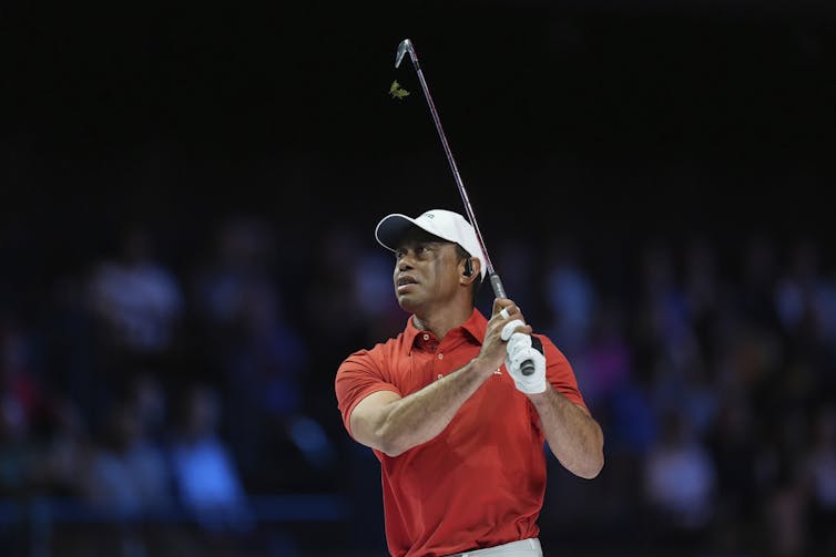 A man wearing a red shirt and white cap swings a golf club.