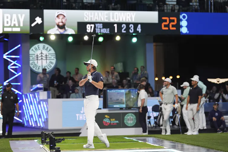 A man in a dark shirt and white pants swings a golf club, others holding clubs stand in the bkacground.