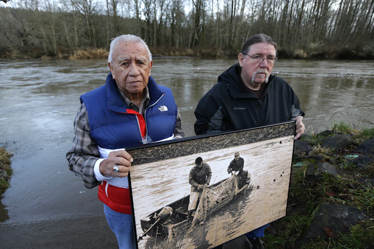 Two men stand in front of a river holding an enlargement of an old photograph.