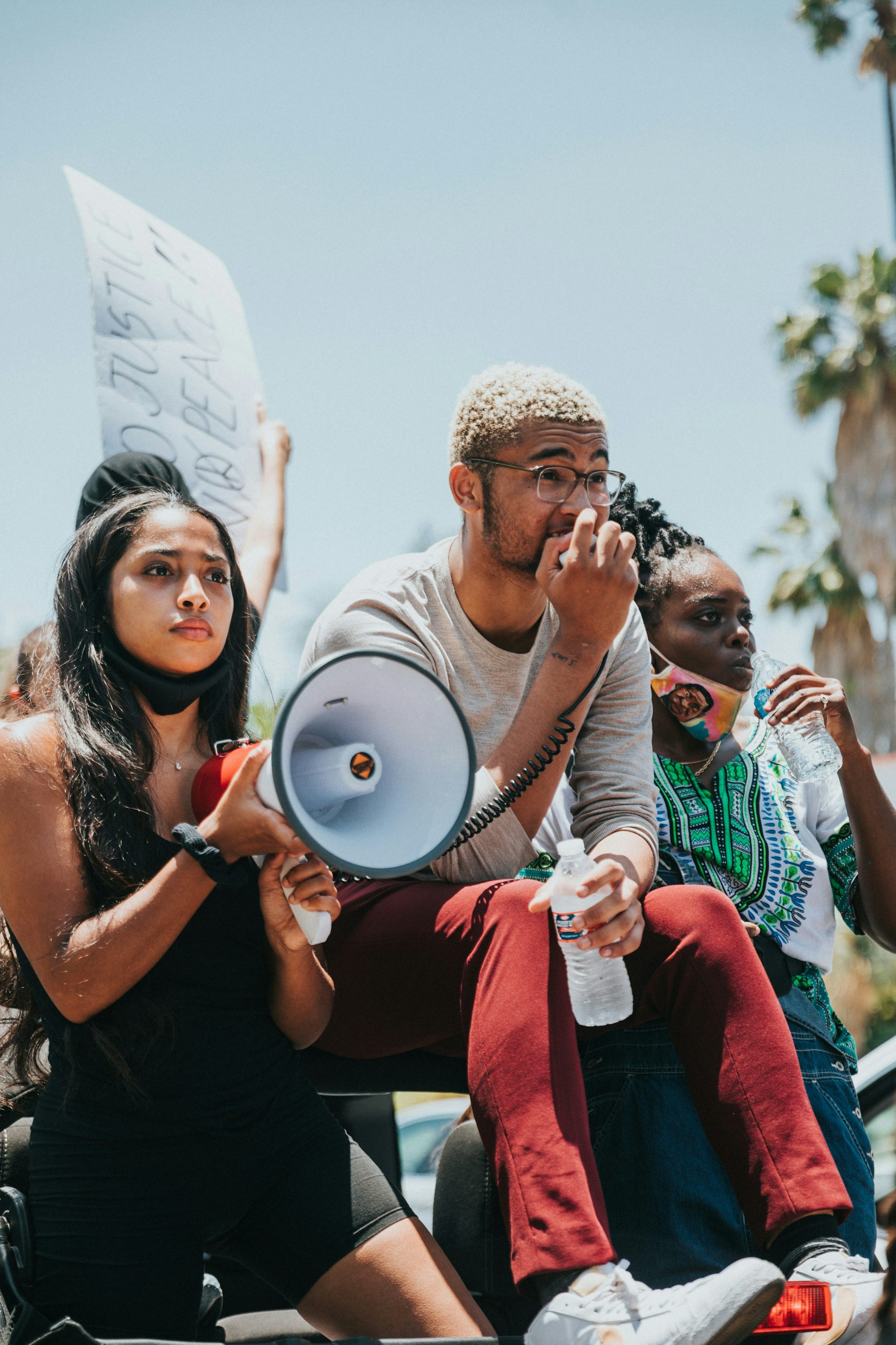 Three people at a demonstration. One person is holding a megaphone while another speaks into the microphone. Another person in the frame is taking a sip of water. Two of the people have masks on, but not covering their faces. 