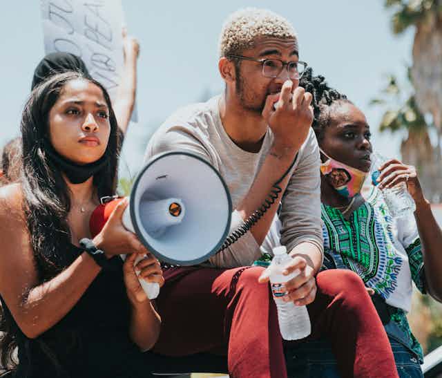 Three people at a demonstration. One person is holding a megaphone while another speaks into the microphone. Another person in the frame is taking a sip of water. Two of the people have masks on, but not covering their faces.
