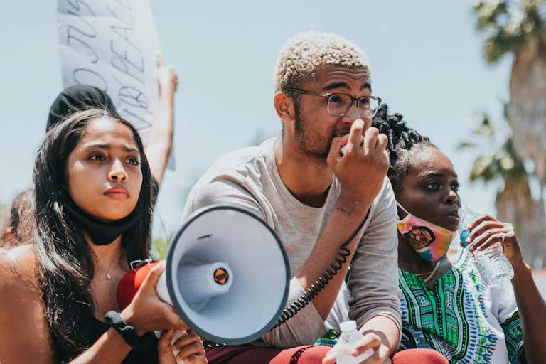 Three people at a demonstration. One person is holding a megaphone while another speaks into the microphone. Another person in the frame is taking a sip of water. Two of the people have masks on, but not covering their faces.