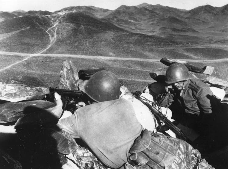 Men in helmet look across an expanse with mountains on the other side.