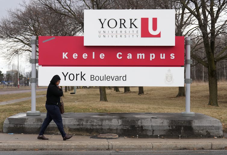Una persona con una chaqueta con capucha pasa por el letrero que dice el campus de York Keele