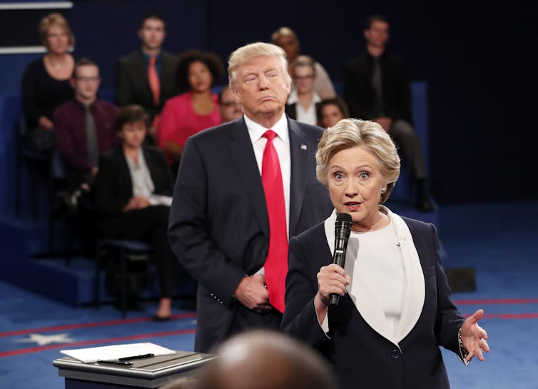 Donald Trump, wearing a red tie, looms over Hillary Clinton as she speaks.
