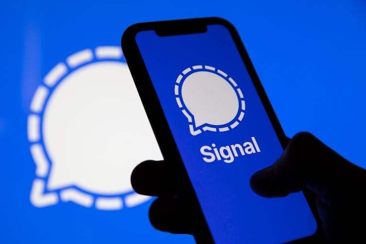 Hand holding a mobile phone displaying the blue and white logo for the Signal app.