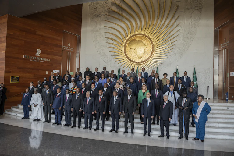 Group of over 40 people, most of them men in suits, standing in rows o n steps in front of a large banner depicting a map of Africa