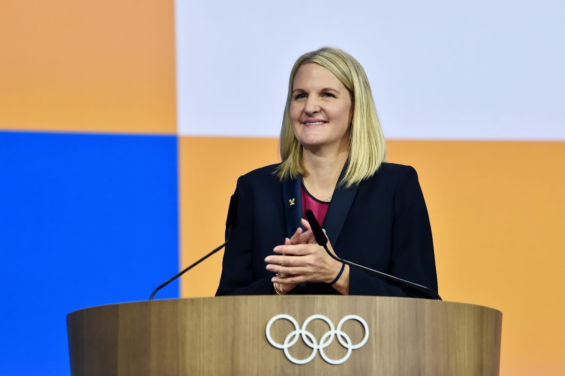 A woman with long blonde hair stands at a podium decorated with the Olympic logo of five rings. She's smiling.