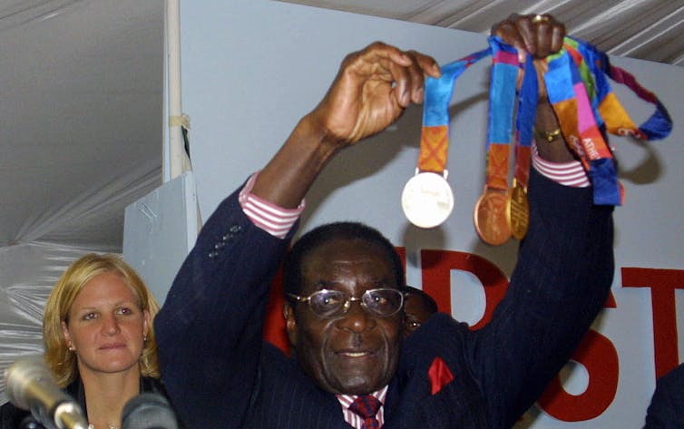 An African man in a suit holds up Olympic medals, a blonde woman standing behind him.