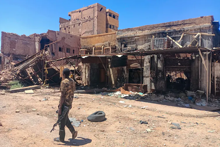 A man with a gun walks down a dirt road with damaged buildings in the backfround