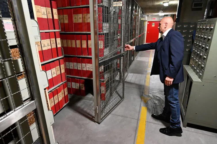 A man in a blue suit swings open a door to storage shelves full of large red books.
